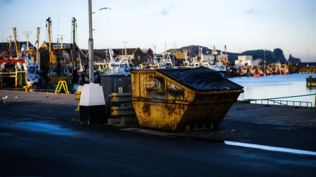A coastal harbor with fishing boats and a yellow dumpster under a clear sky. Stocking a dumpster rental business: inventory and suppliers